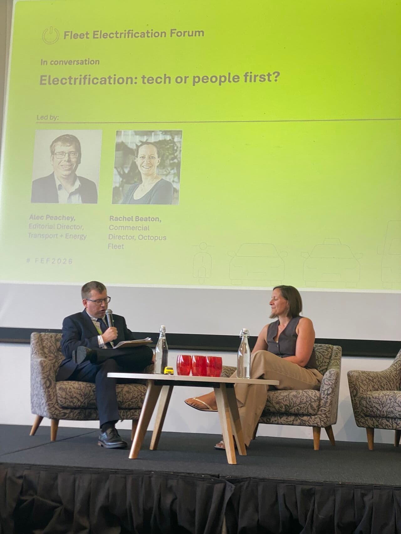 A man and a woman sitting on stage discussing at the Fleet Electrification Forum. A large screen behind them shows the event's title and speakers' names.