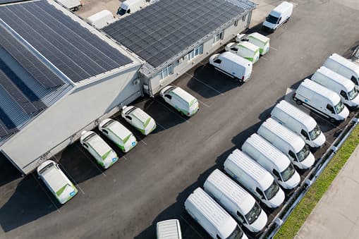 Aerial view of a fleet of white delivery vans parked around a building with solar panels on its roof, under a clear sky.