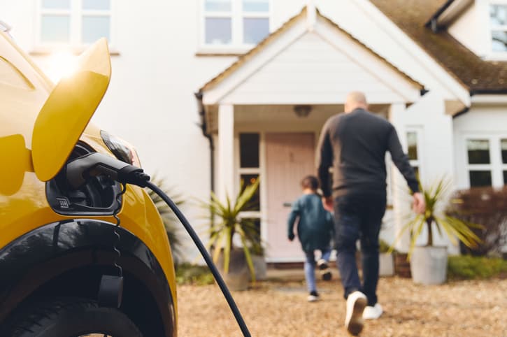 Yellow electric car charging in a driveway; a man and child walking towards a house with a pink door.